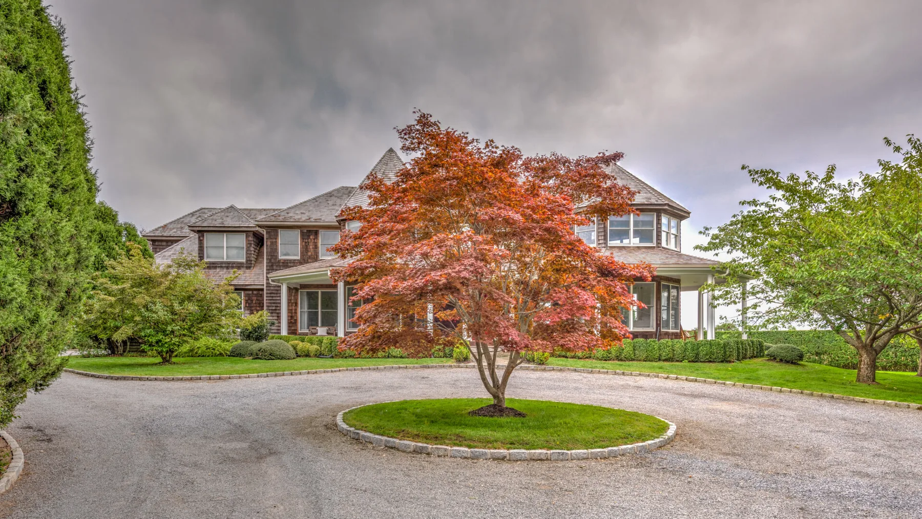 Una casa grande se alza detrás de una entrada circular y un árbol de hojas rojas.