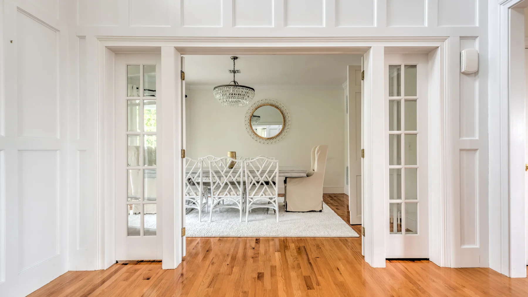 Un comedor luminoso con asientos blancos, una lámpara de araña y un espejo redondo.