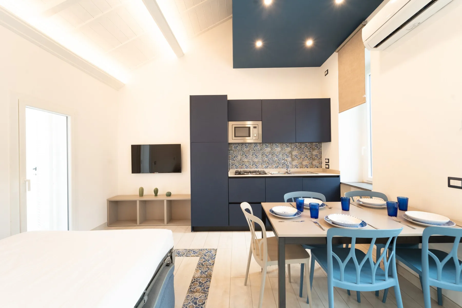 A bright open-plan room with a compact kitchen, dining table, and bed.