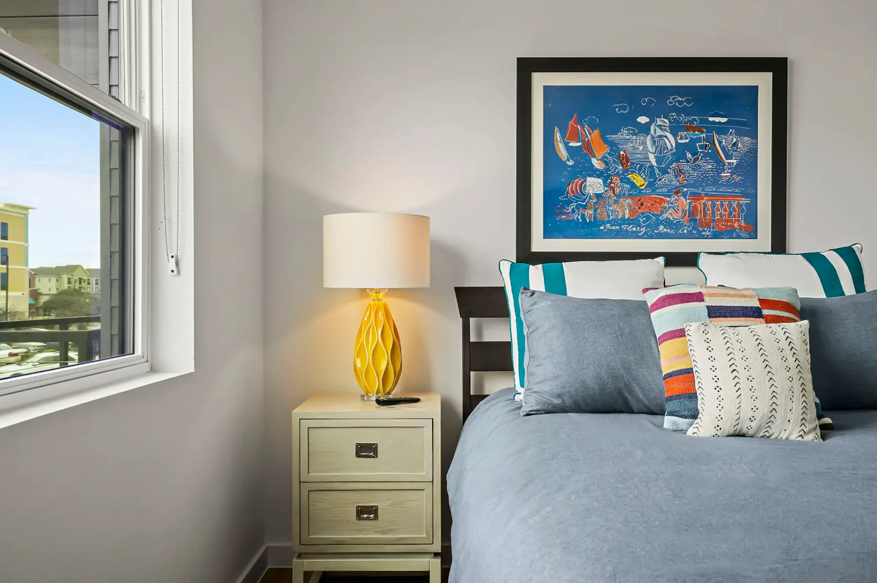 Rincón de un dormitorio con una cama, cojines de colores, una lámpara amarilla sobre una mesa de noche y un cuadro abstracto enmarcado en la pared.