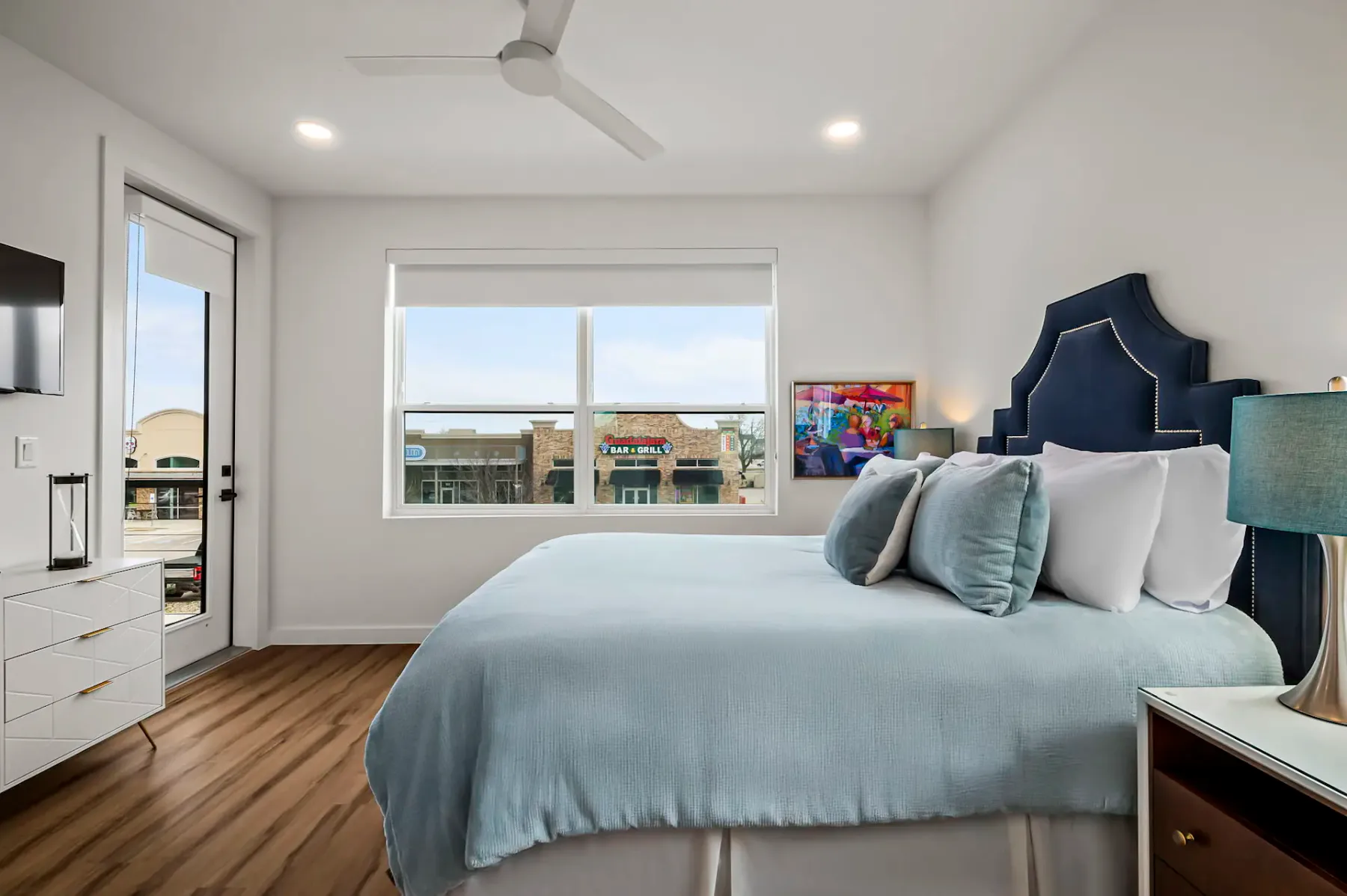 Dormitorio moderno con una cama grande, ropa de cama azul y blanca, cabecera oscura, piso de madera y una ventana con vistas a edificios. Cuenta con ventilador de techo y lámparas.