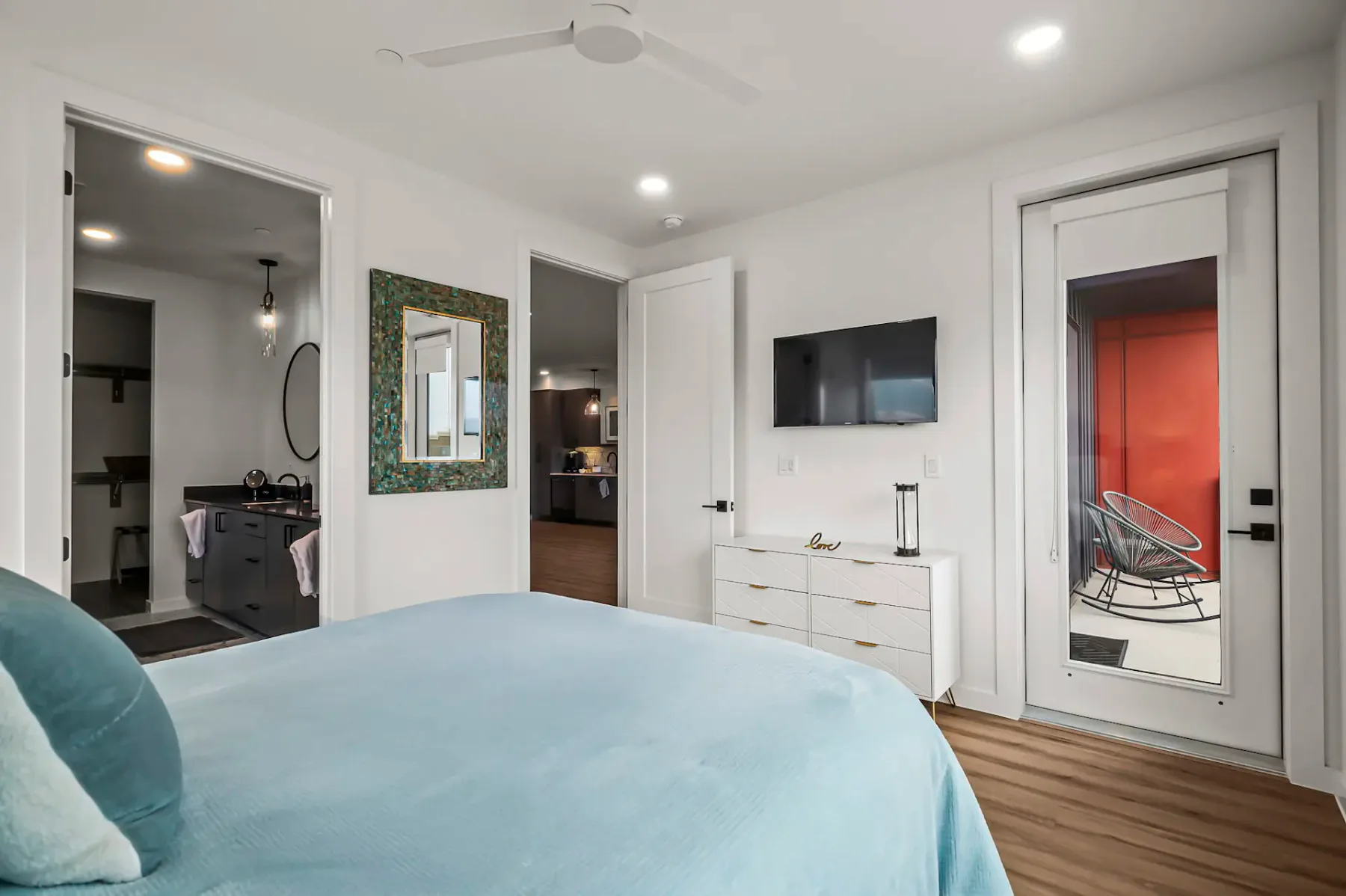 Dormitorio moderno con una colcha azul, televisor montado en la pared, cómoda blanca, puerta de vidrio y puertas abiertas que dan a un baño y a otra habitación.