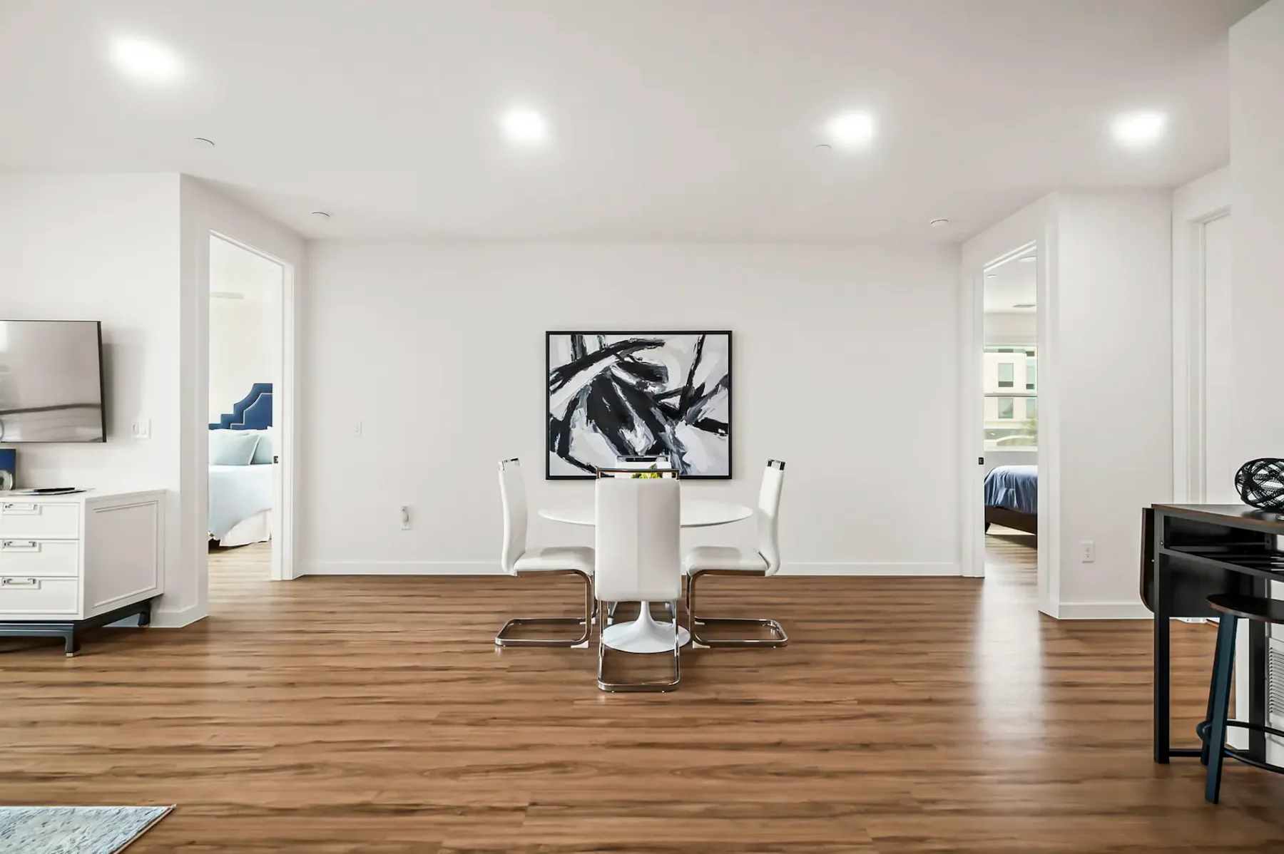 Comedor moderno con una pequeña mesa redonda y cuatro sillas blancas sobre piso de madera, con arte abstracto en la pared y puertas abiertas contiguas.