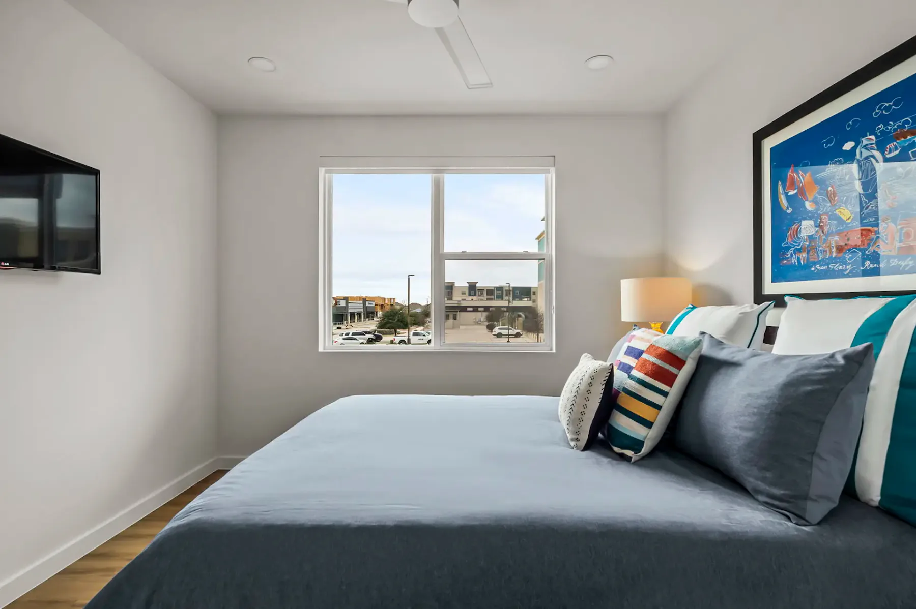 Dormitorio minimalista con una cama grande, cojines de colores, arte en la pared, una ventana con vista a la ciudad y un televisor montado en la pared.