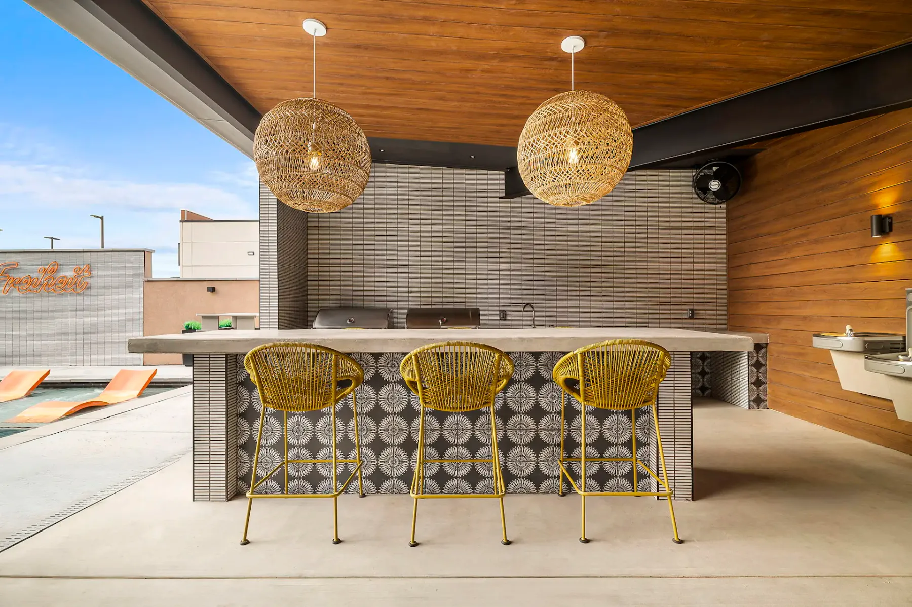 Cocina al aire libre con una barra revestida de azulejos, tres taburetes amarillos y dos grandes lámparas colgantes de material tejido bajo un techo de madera.
