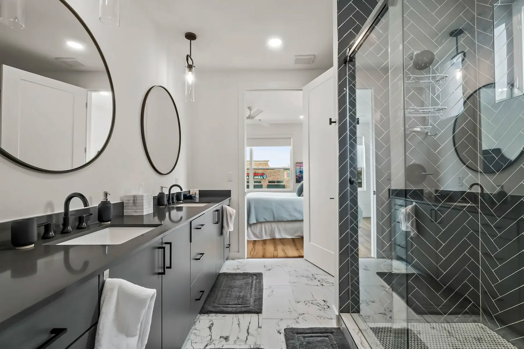 Baño moderno con dos lavabos, espejos grandes, gabinetes grises y una ducha con cerramiento de vidrio. Al fondo se ve un dormitorio a través de una puerta abierta.