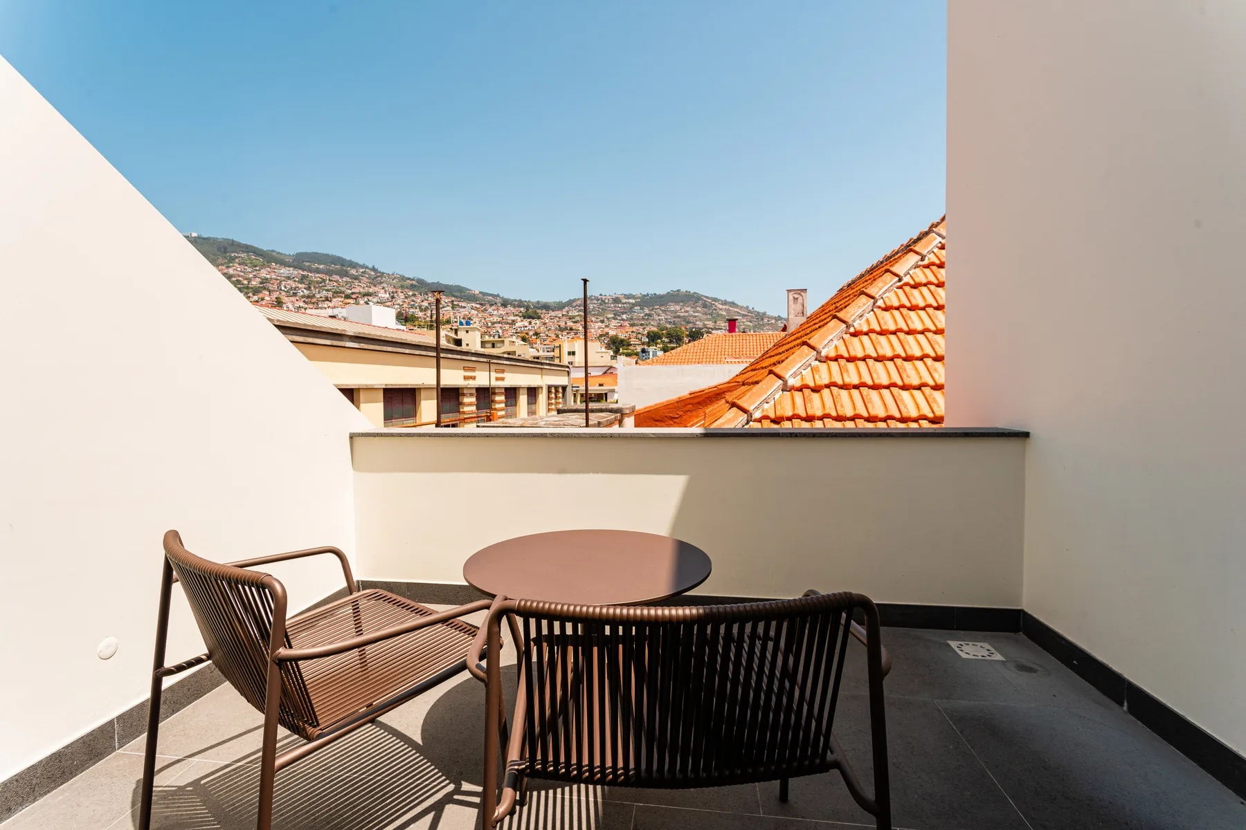 A rooftop terrace has a round table and seating overlooking nearby rooftops and hills.