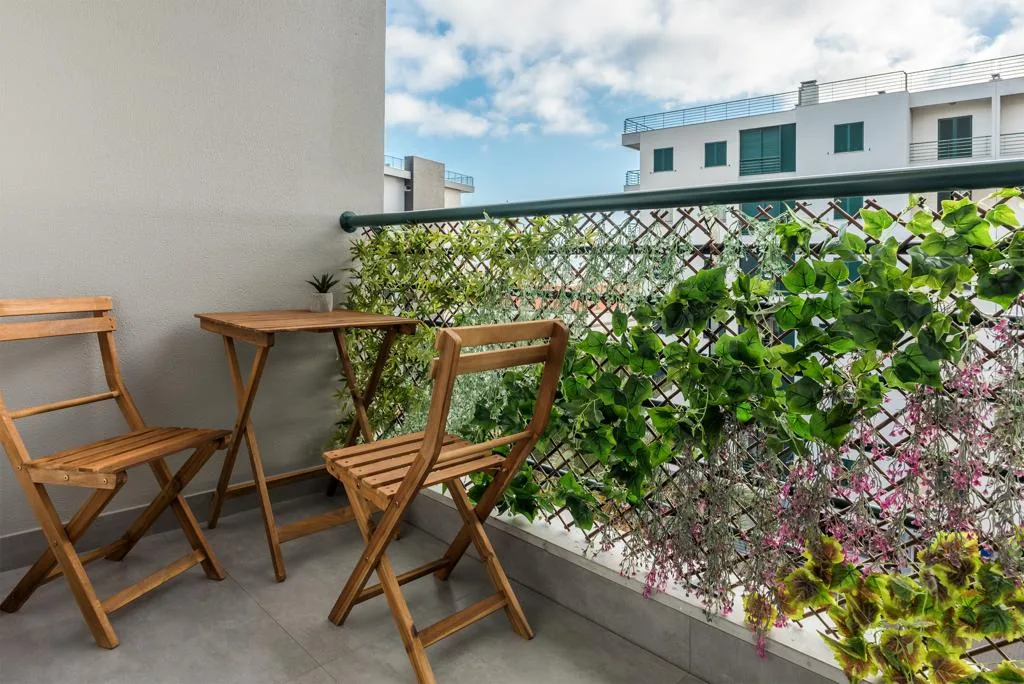 A balcony seating area with wooden chairs and a small table overlooks leafy plants and nearby buildings.