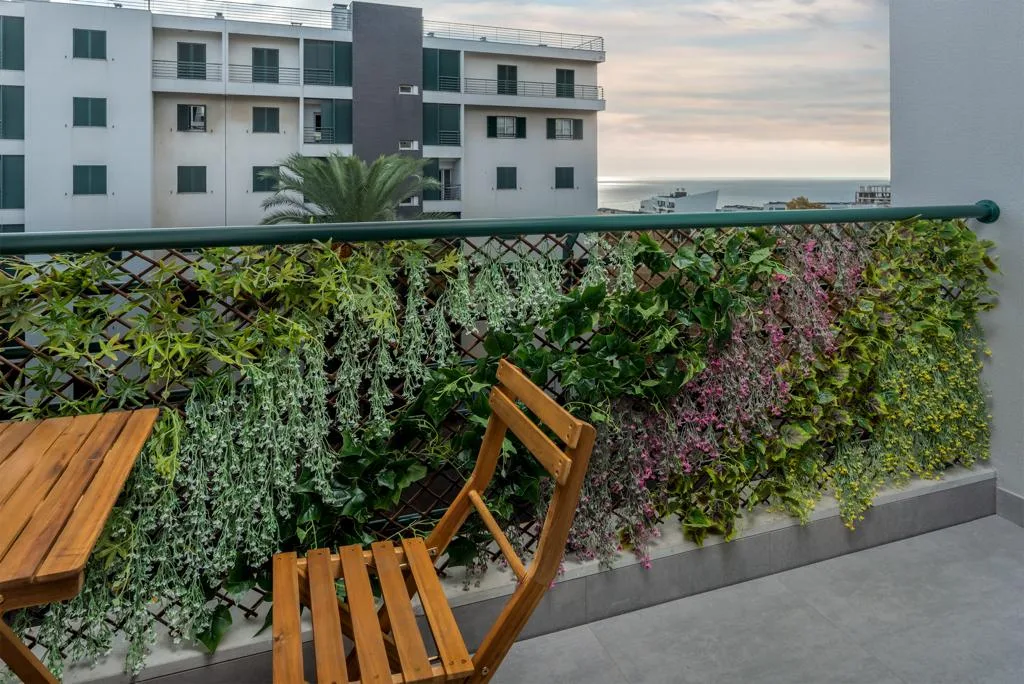 A balcony with wooden seating, a leafy green screen, and a view of nearby buildings and the sea.
