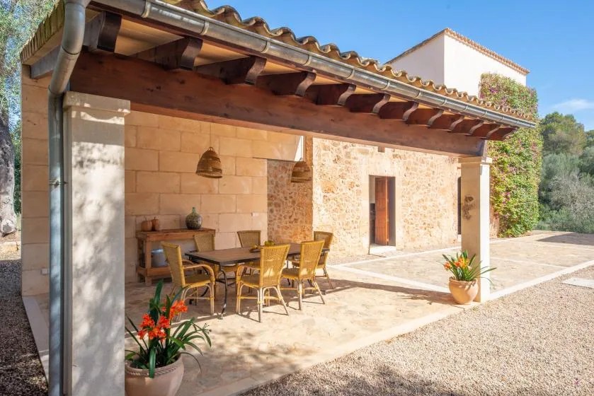 A stone patio with outdoor seating sits beneath a shaded roof beside the house.