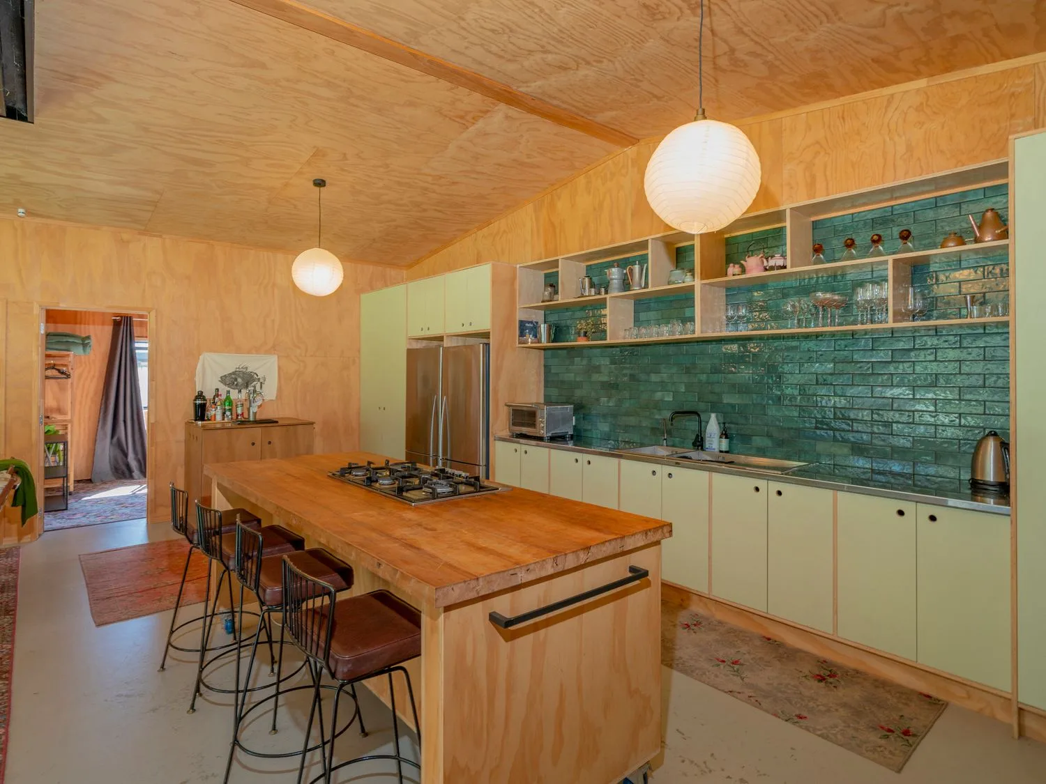 Una cocina con una isla de madera, taburetes de barra, un salpicadero de azulejos verdes, armarios y lámparas colgantes en The Pumpkin Shed, en Kuaotunu
