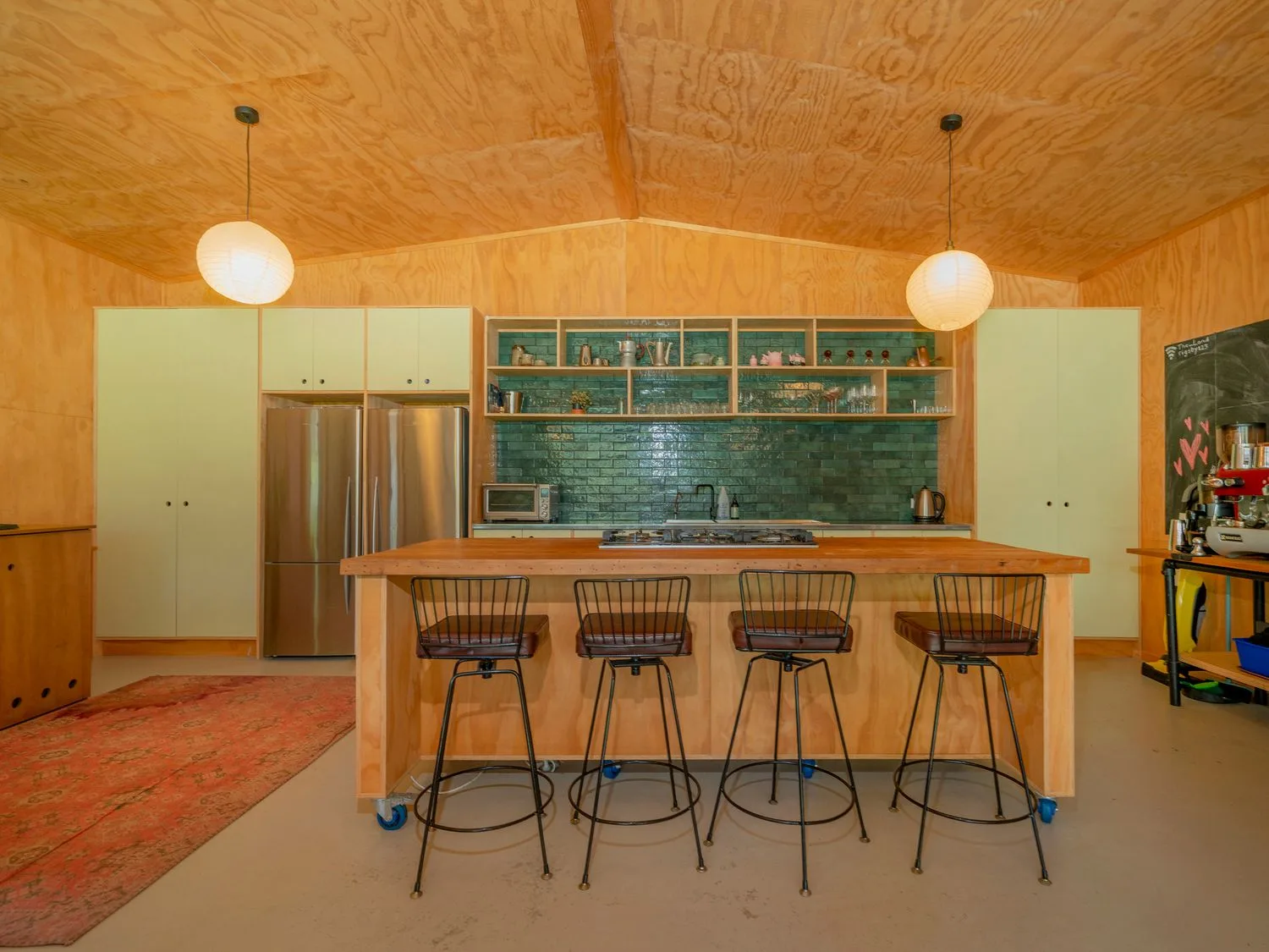 Una cocina con una isla de madera y cuatro taburetes de metal, un salpicadero de azulejos verdes y armarios blancos en The Pumpkin Shed - Kuaotunu Holiday Home, en Kuaotunu