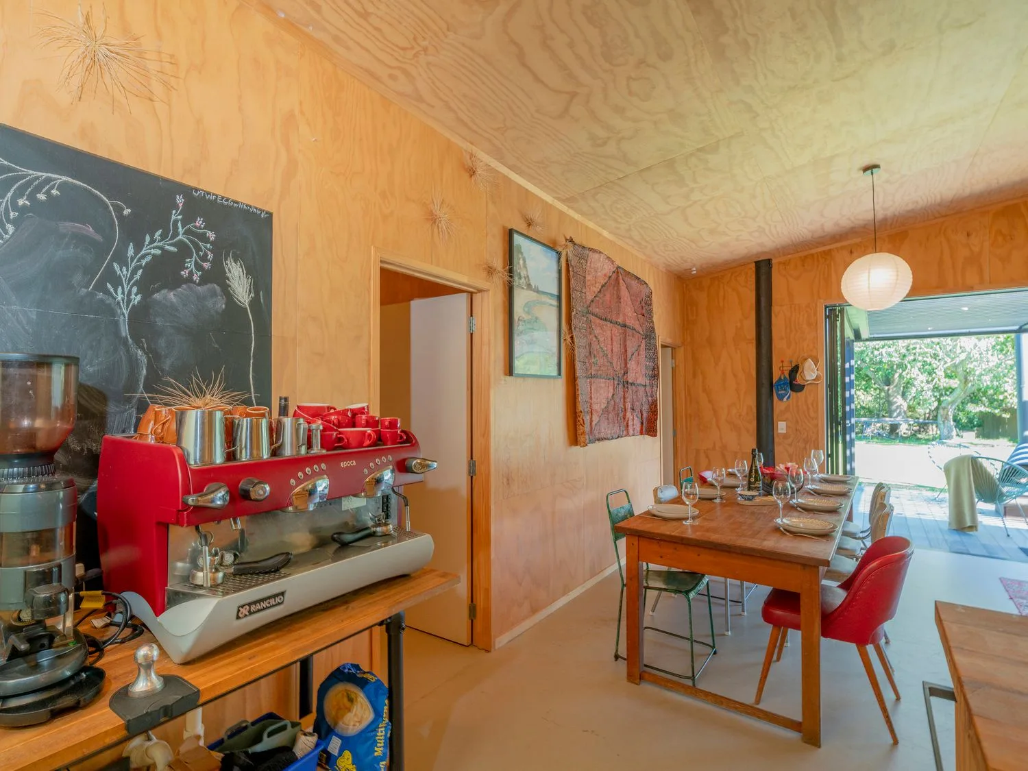 Un comedor con una mesa de madera puesta con platos y vasos y una cafetera sobre una encimera, en una habitación con paredes de madera y una puerta abierta en The Pumpkin Shed Kuaotunu