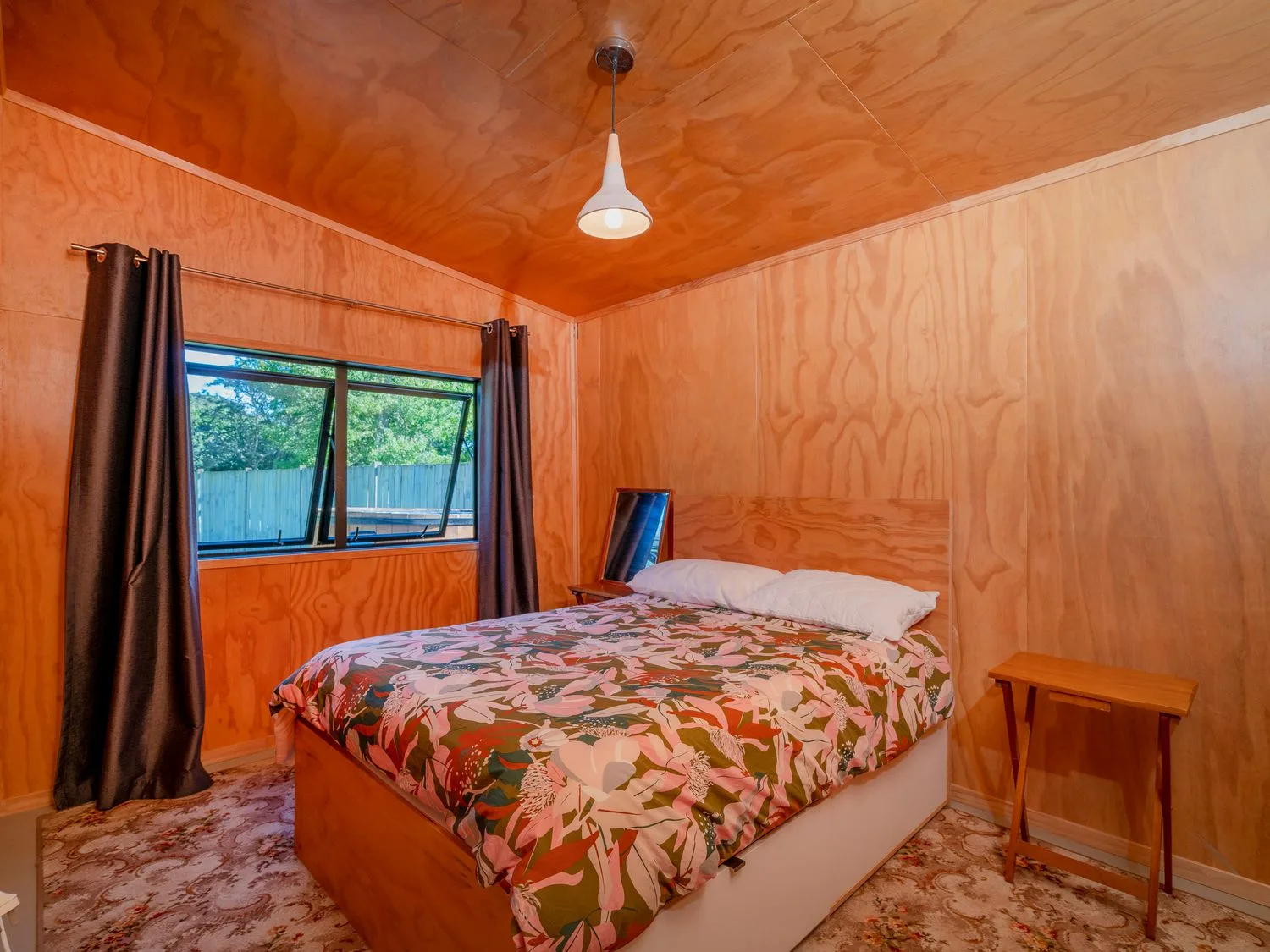 Un dormitorio con una cama con ropa de cama de estampado floral, una ventana con cortinas, una lámpara colgante y una pequeña mesa de madera en The Pumpkin Shed Kuaotunu Holiday Home, en Kuaotunu