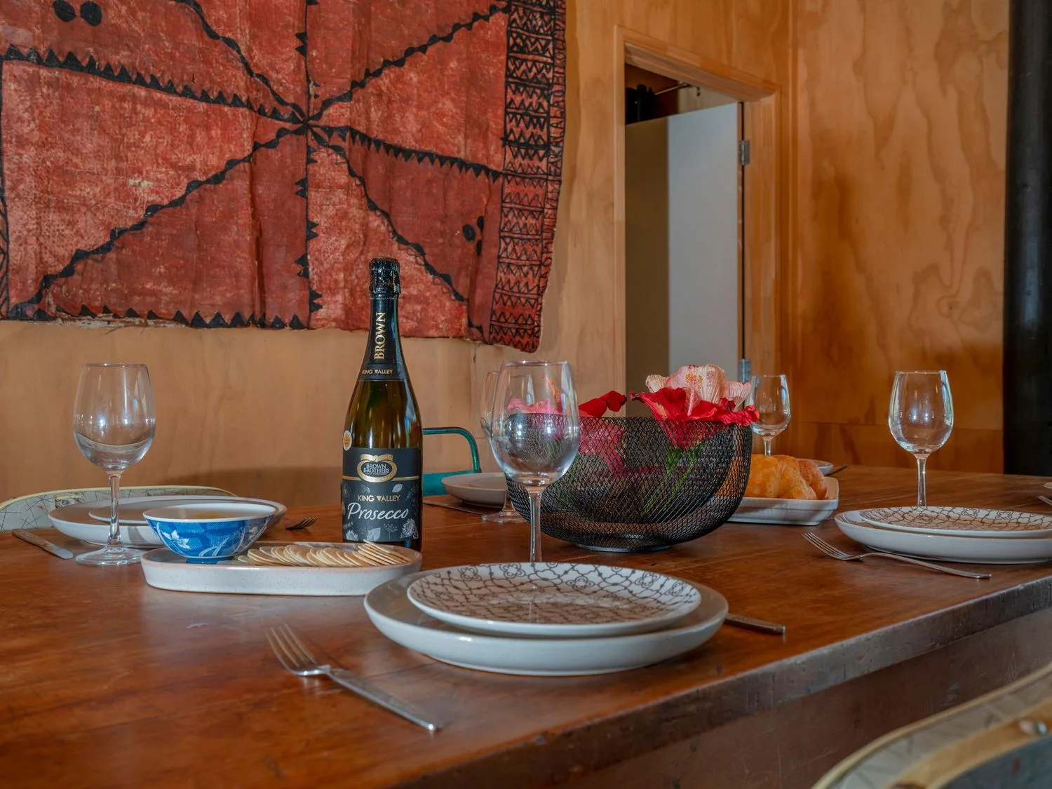 Una mesa de comedor puesta con platos, vasos, una botella de prosecco, galletas saladas y un cuenco con flores en una habitación con paredes de madera en The Pumpkin Shed - Kuaotunu Holiday Home, en Kuaotunu