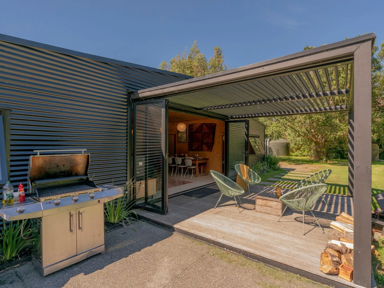 Un patio exterior con una barbacoa y asientos bajo una pérgola junto a una casa con una puerta abierta en The Pumpkin Shed - Kuaotunu Holiday Home, en Kuaotunu