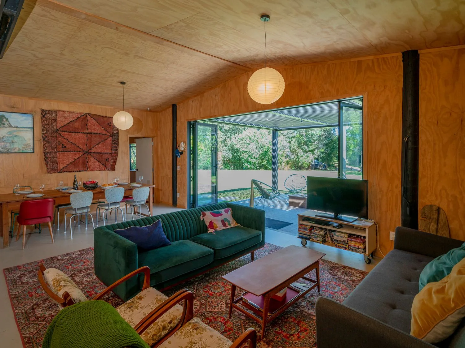 Una sala con sofás verdes y grises, una mesa de centro de madera y un televisor junto a un comedor con una mesa y sillas en The Pumpkin Shed - Kuaotunu Holiday Home, en Kuaotunu