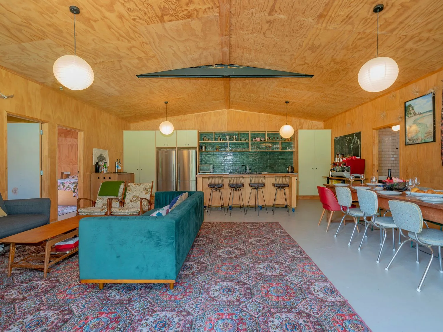 Una zona abierta de sala y comedor con sofás, sillas, una alfombra estampada, una mesa de comedor con sillas y una cocina con taburetes de barra en The Pumpkin Shed - Kuaotunu Holiday Home, en Kuaotunu
