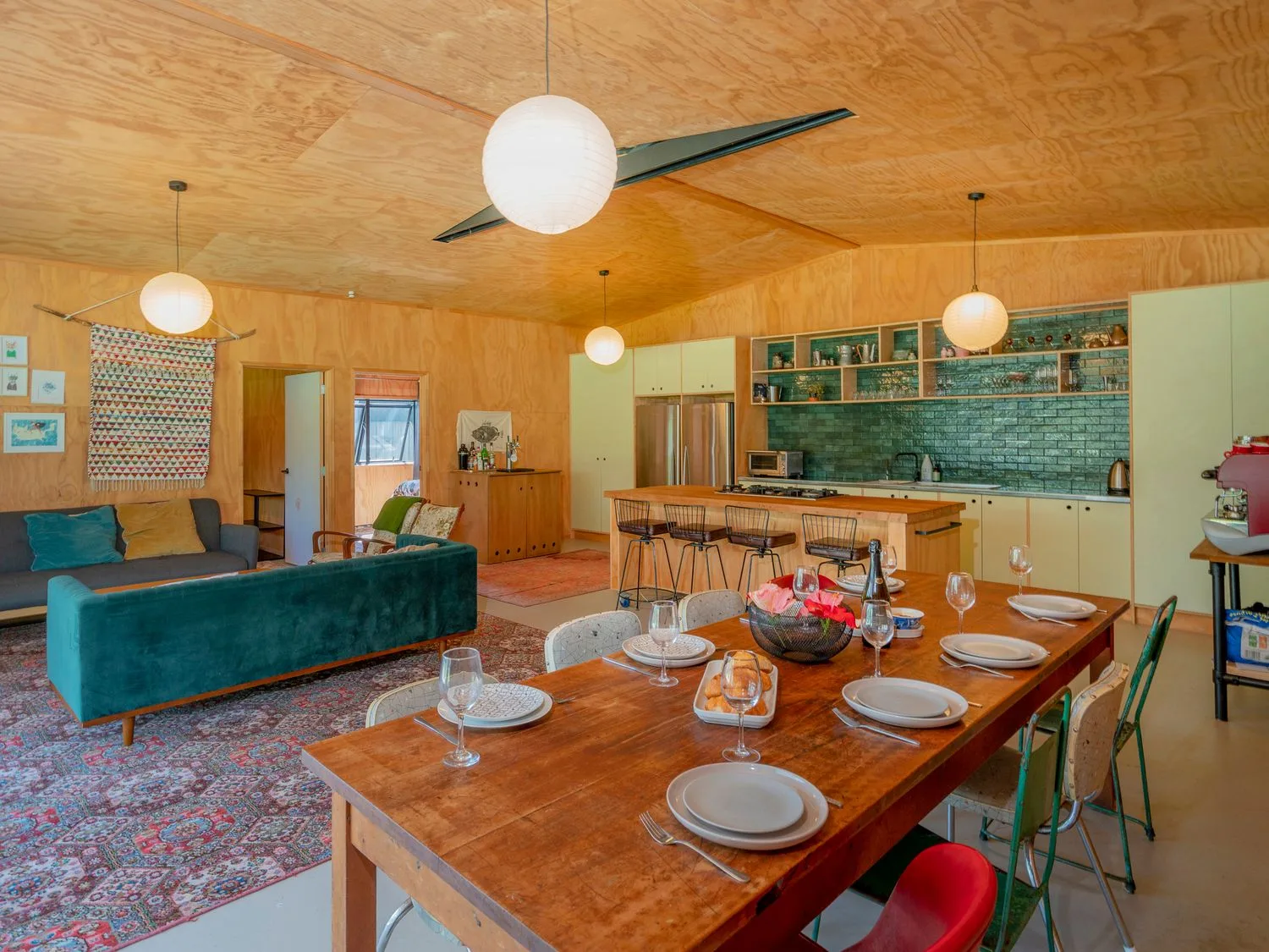Una zona de sala y comedor de planta abierta con una mesa de comedor de madera preparada para seis personas cerca de una cocina con isla y taburetes de barra en The Pumpkin Shed, en Kuaotunu