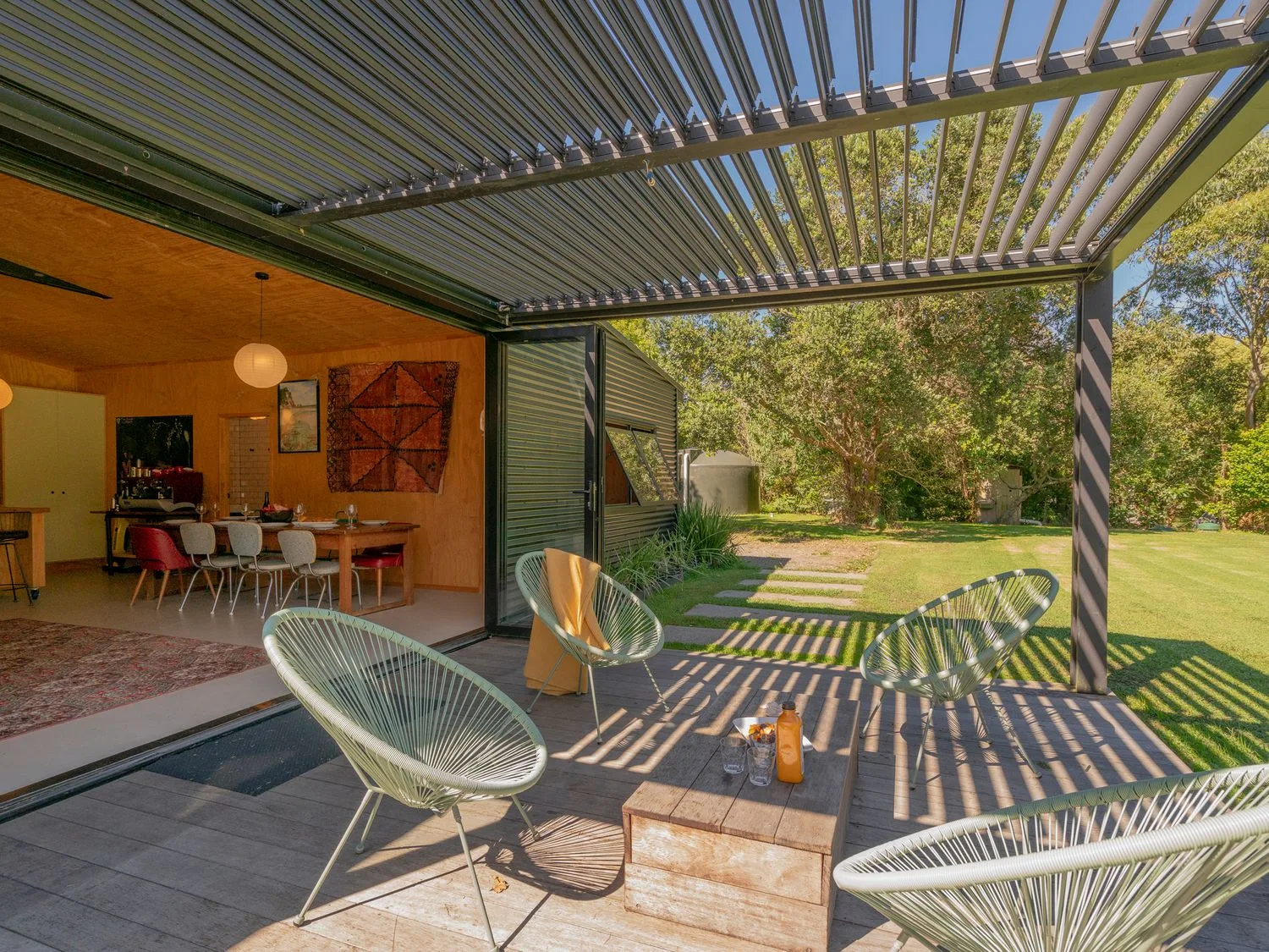 Un patio exterior con una pérgola de listones se abre a un comedor amueblado y un jardín.