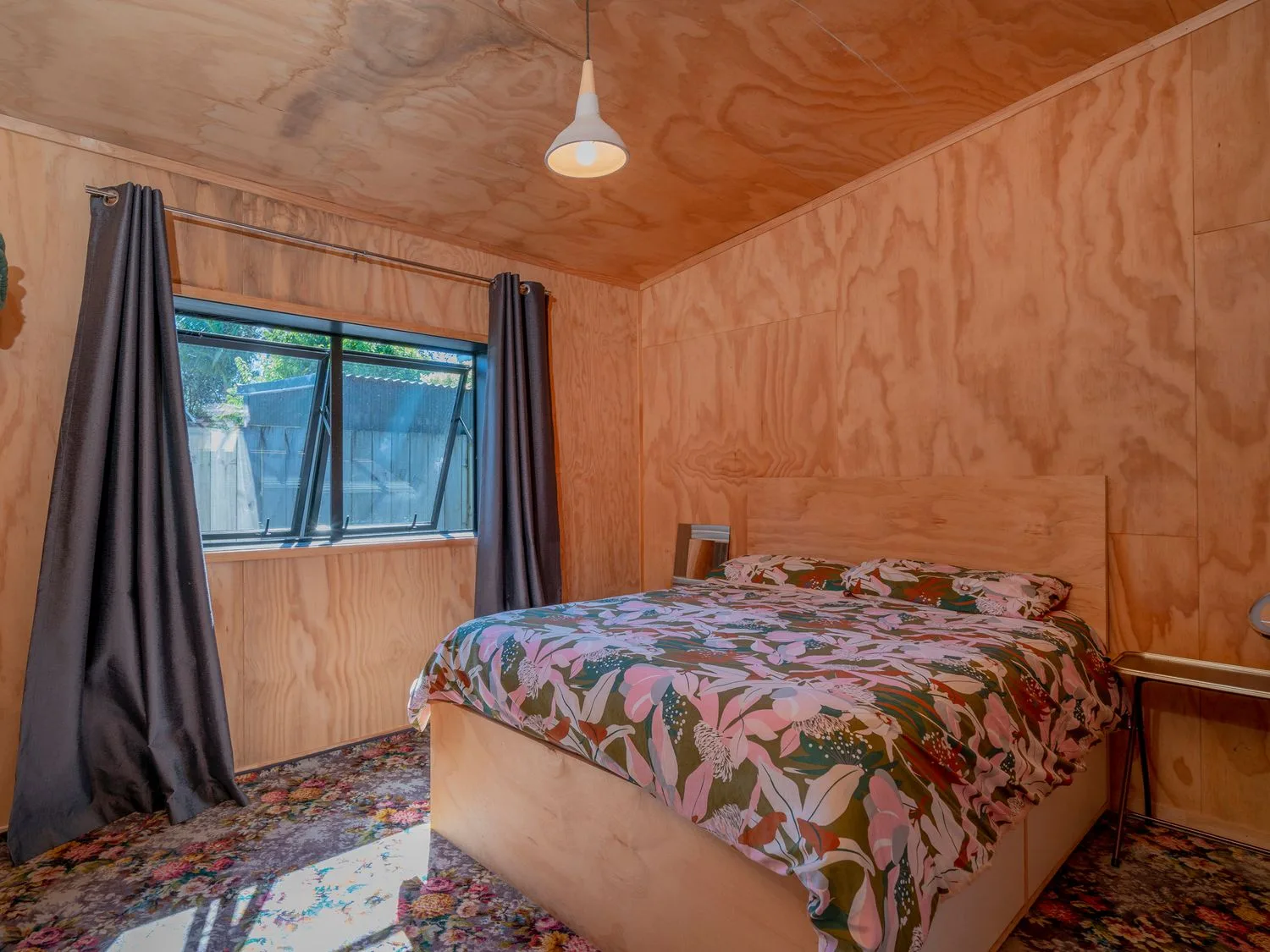 Un dormitorio con una cama con ropa de cama de estampado floral, una ventana con cortinas oscuras y paredes de madera en The Pumpkin Shed, en Kuaotunu