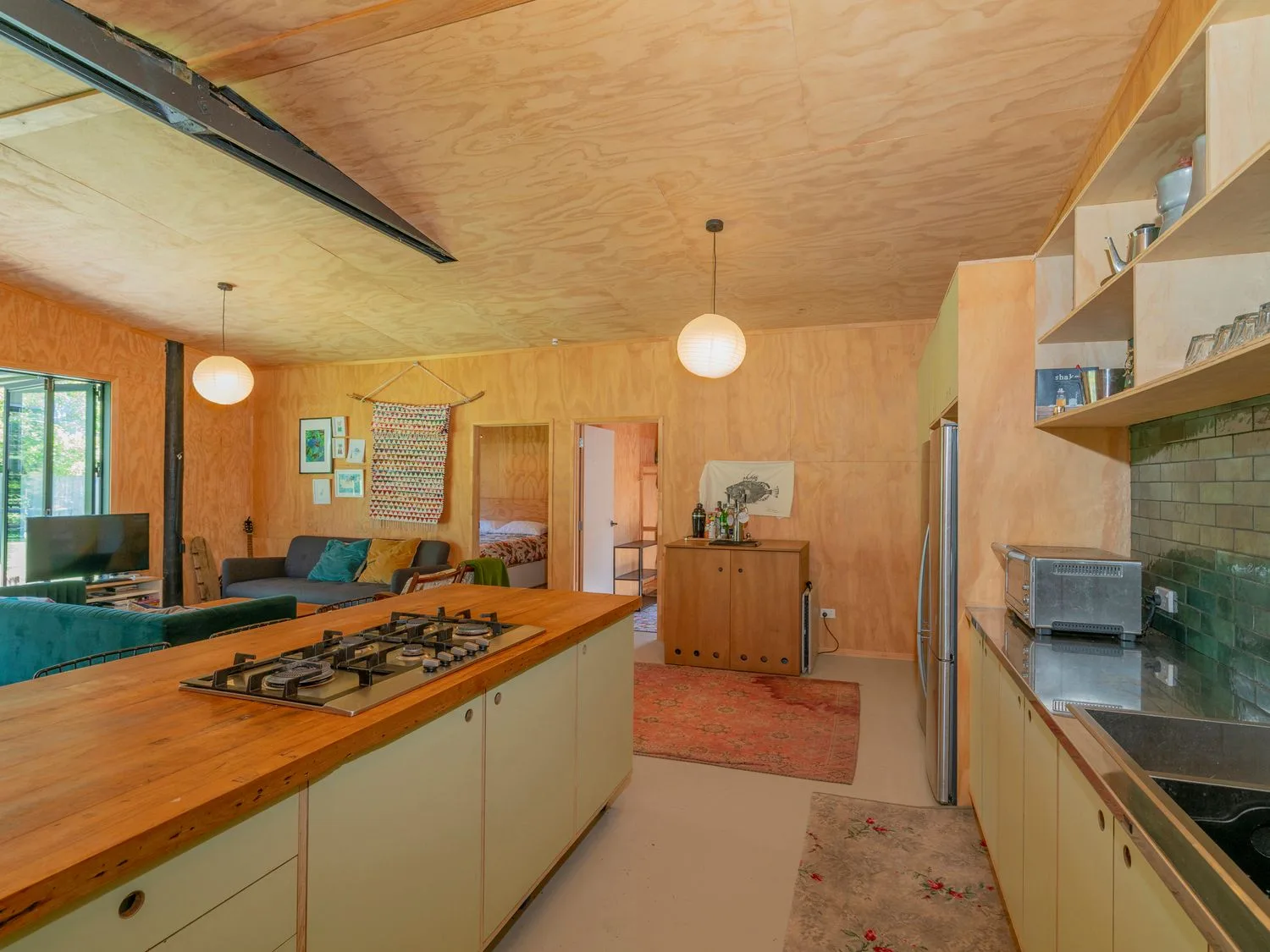 Una cocina y una sala de planta abierta con una isla de madera y una placa de cocina, un sofá, un televisor y decoraciones en las paredes en The Pumpkin Shed Kuaotunu Holiday Home, en Kuaotunu