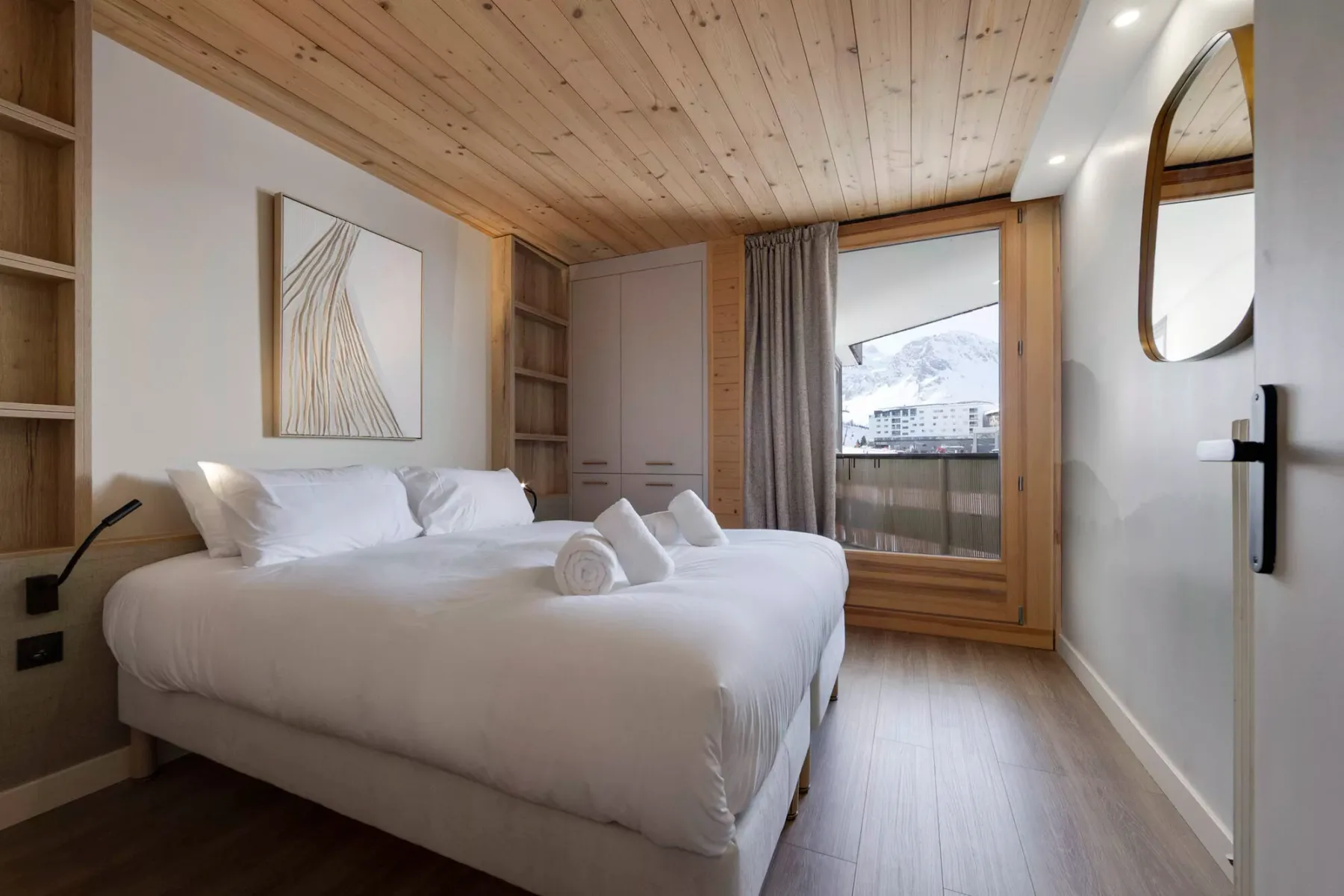 A bright bedroom with a neatly made bed and a window overlooking snowy mountains.