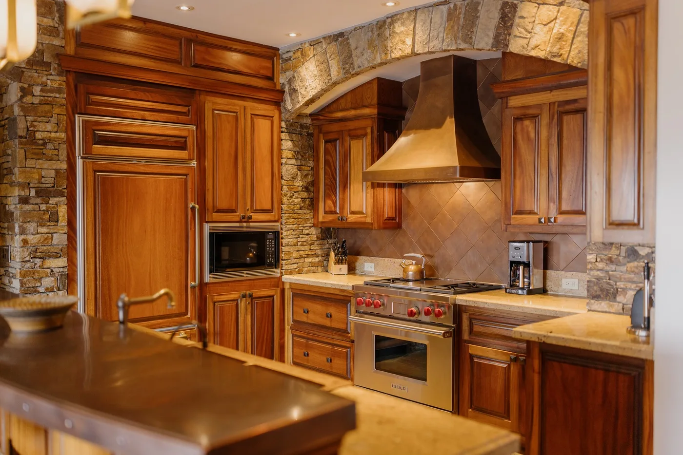A kitchen with wooden cabinetry, stone walls, and a central cooking range.