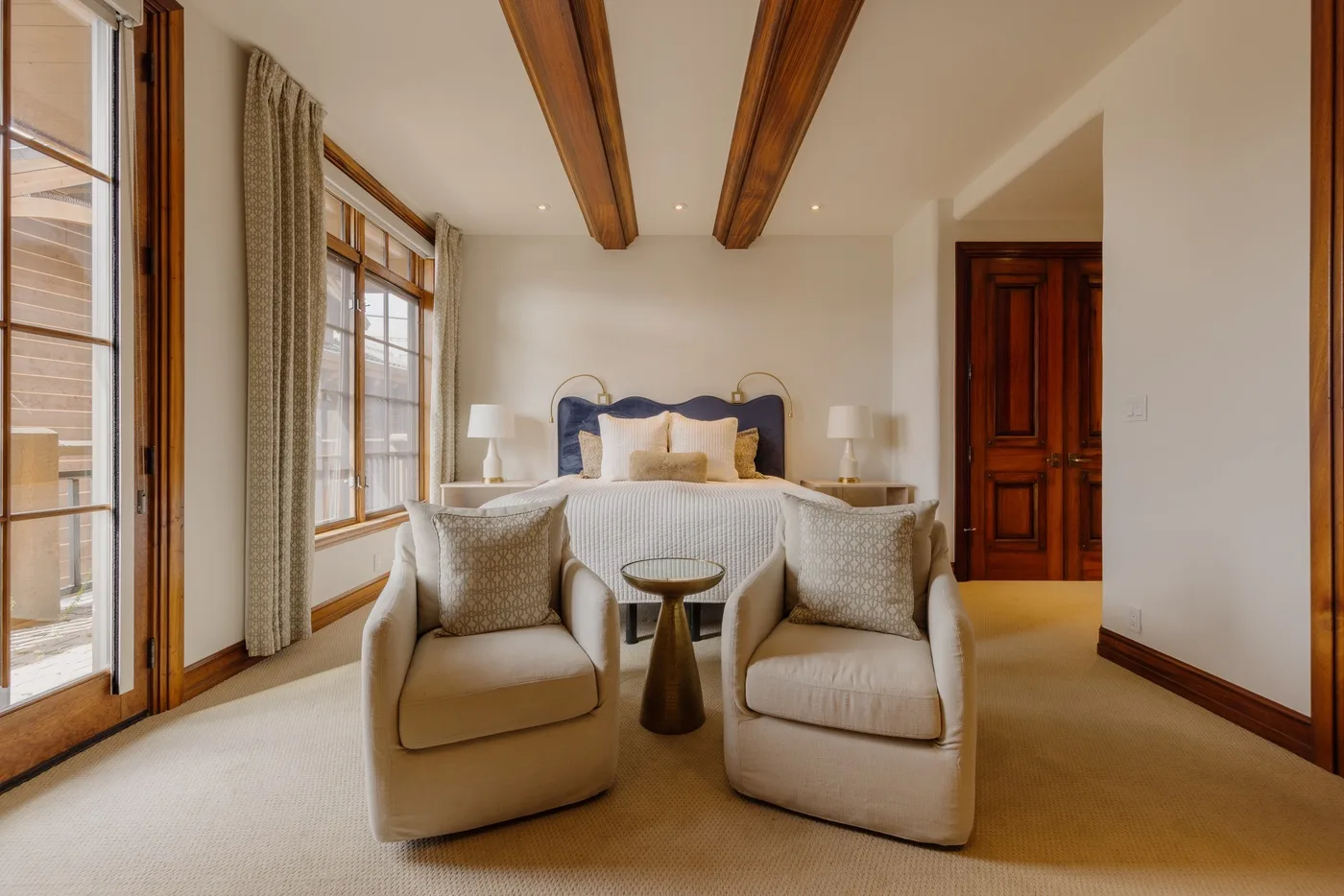 A bedroom with two upholstered chairs facing a bed beside large windows.