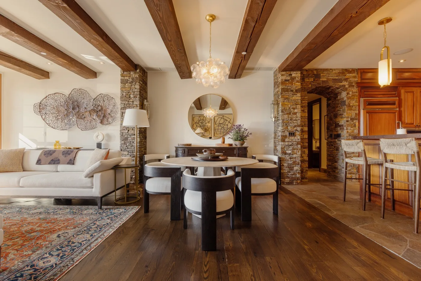 An open dining and living area features a round table beneath exposed wooden beams.