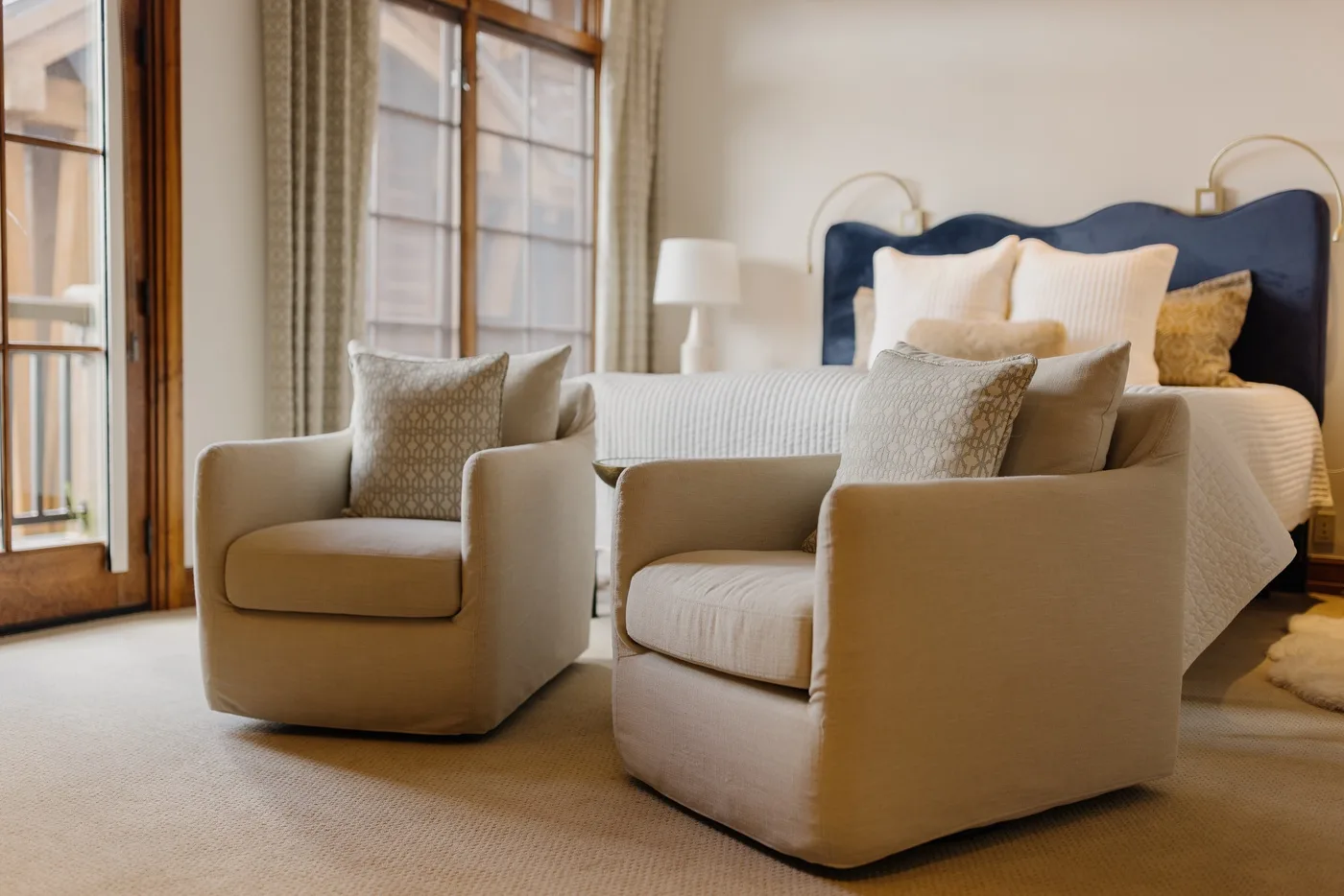 A bedroom with a bed and two upholstered armchairs beside tall windows.