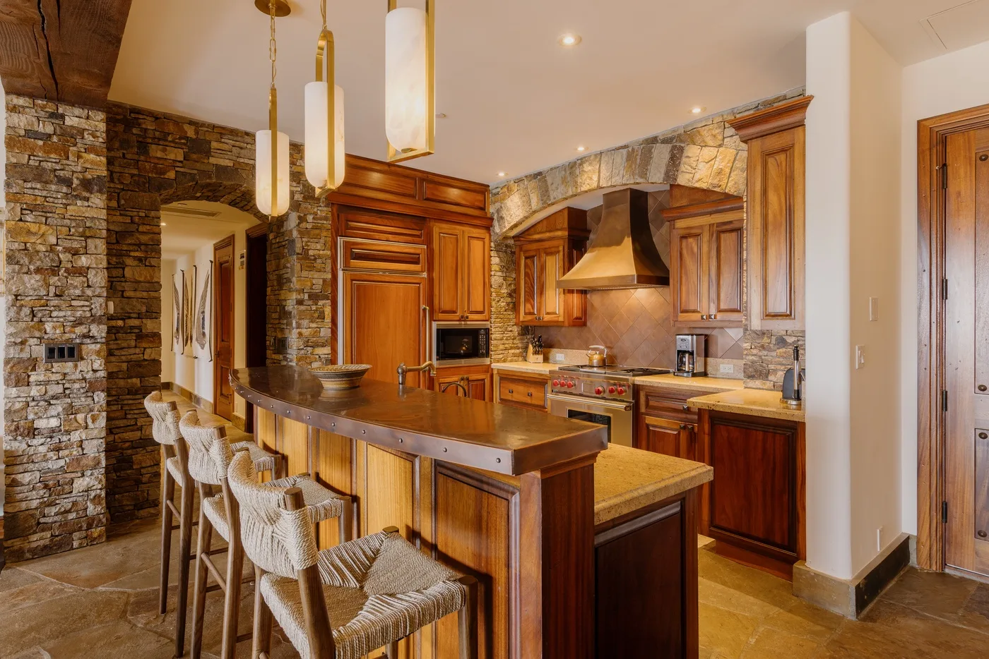 A warm-toned kitchen with wooden cabinetry, stone walls, and a long breakfast bar.