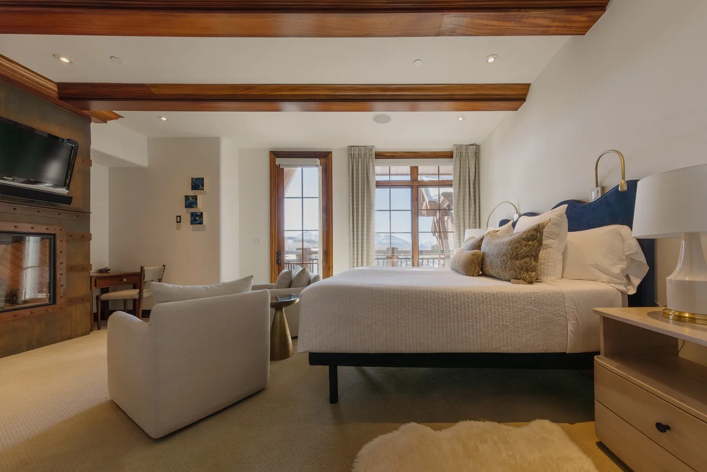 A bedroom with a large bed, seating, and windows beneath exposed wooden beams.