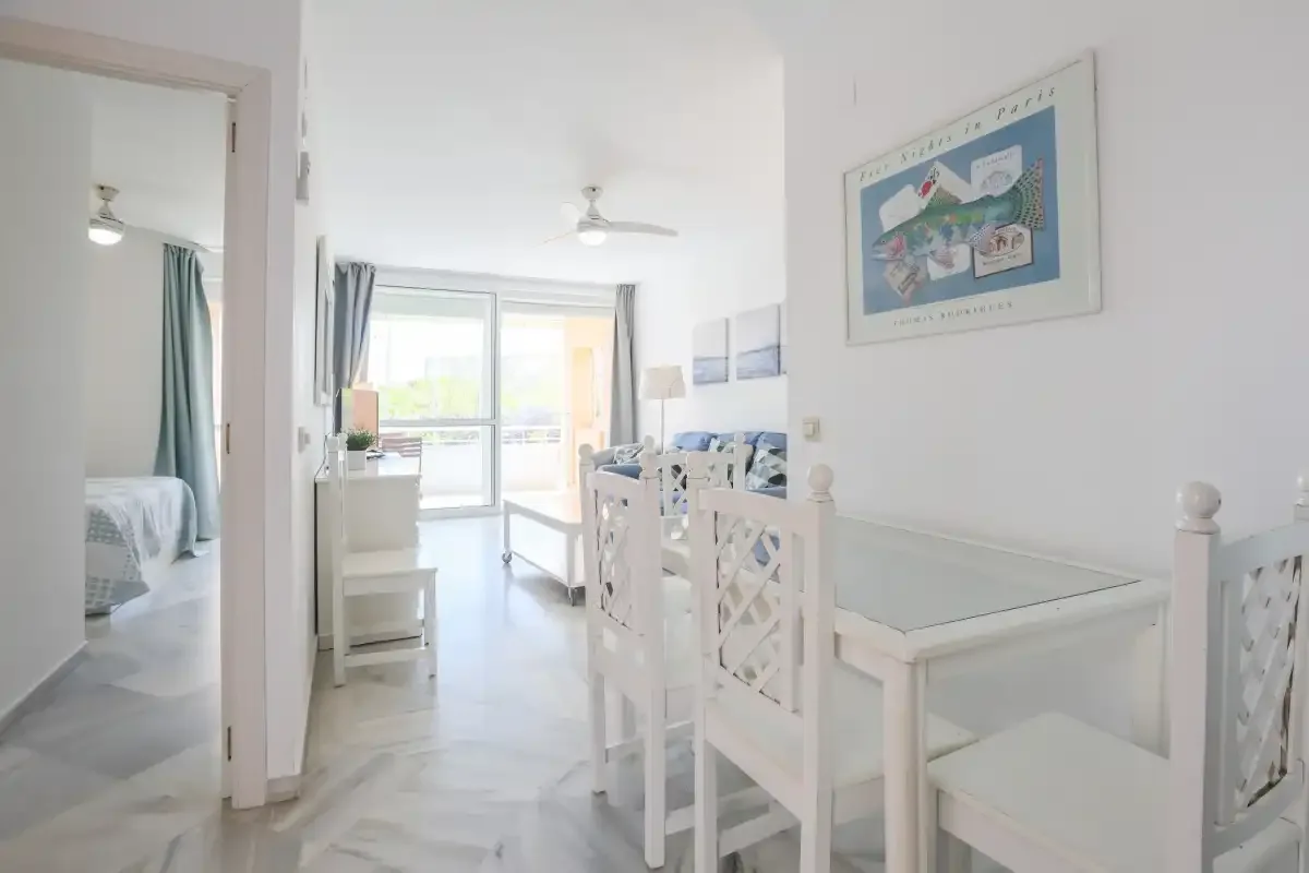 A bright dining area with white seating and a table beside a sunlit balcony.