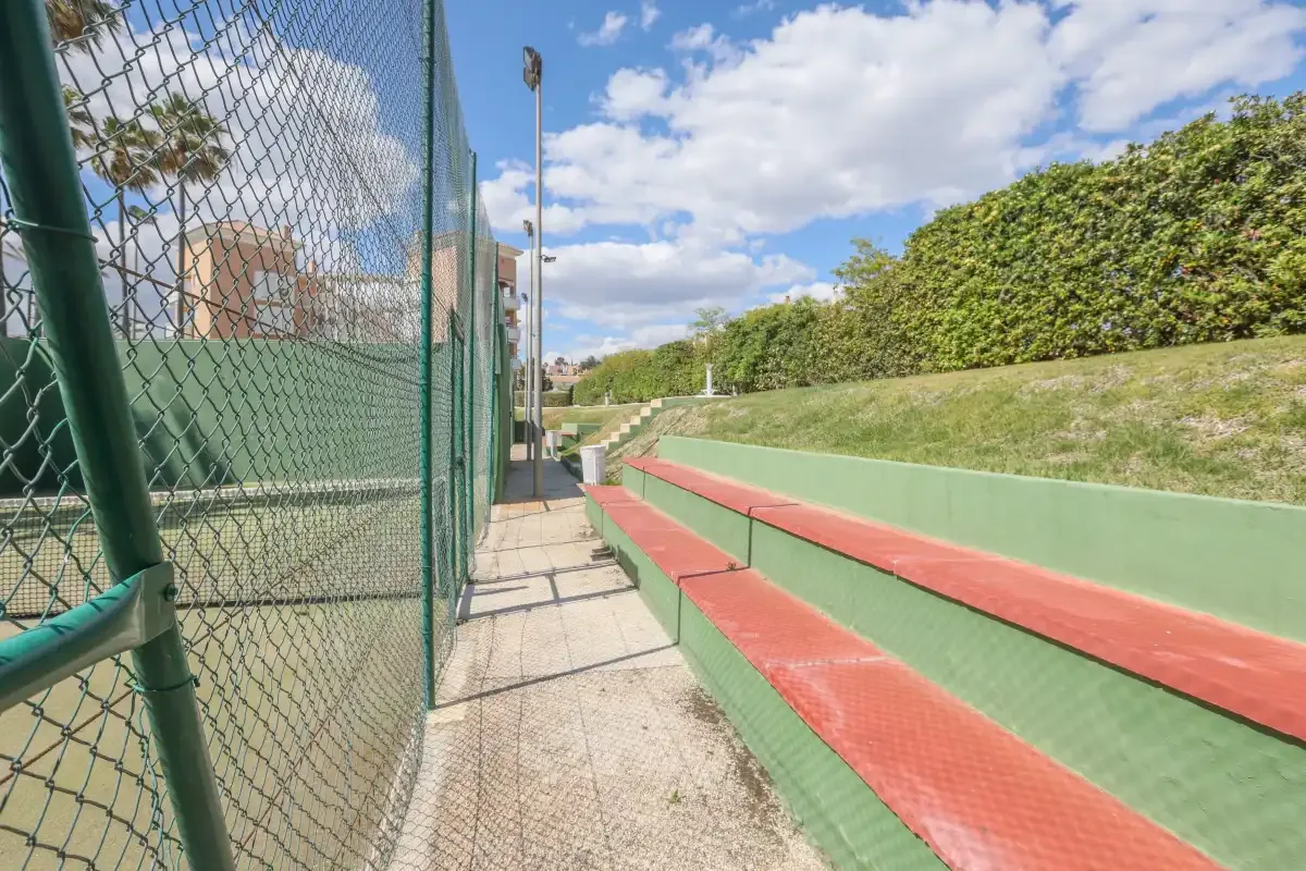 A fenced outdoor sports court has green and red stepped seating beside a hedge.