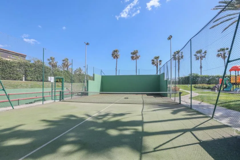 An outdoor court with a net and tall green wall, enclosed by fencing.