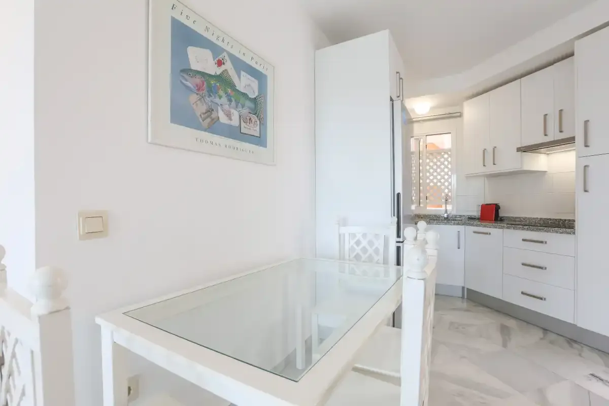 A dining area with a glass-top table beside a white kitchen.