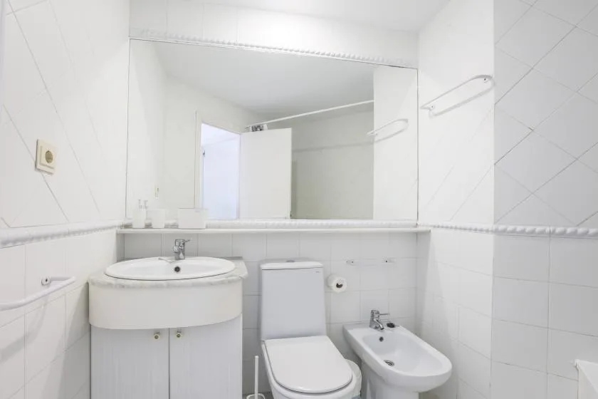 A bright white-tiled bathroom with a sink, toilet, and bidet.