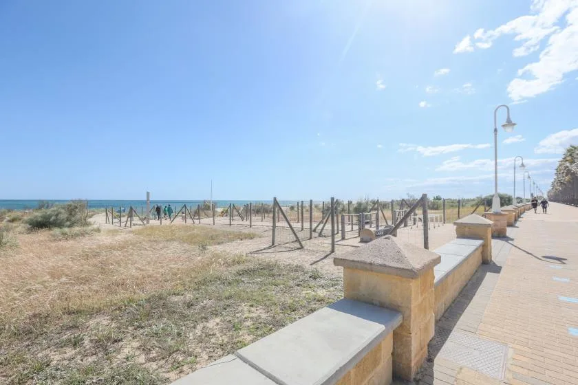 A seaside walkway runs beside a sandy beach and low stone railing.