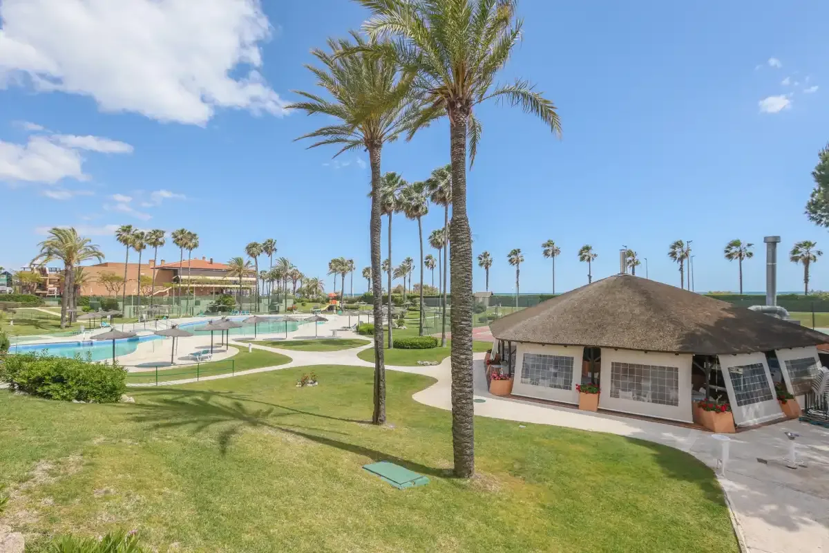 A landscaped outdoor pool area with palm trees, lawns, and a small pavilion.