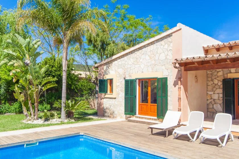 Una casa de piedra da a una terraza junto a la piscina, con tumbonas blancas y contraventanas verdes.