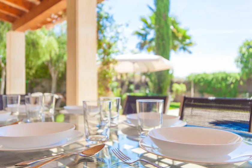 Una mesa de comedor al aire libre está puesta con platos y vasos bajo un patio cubierto.