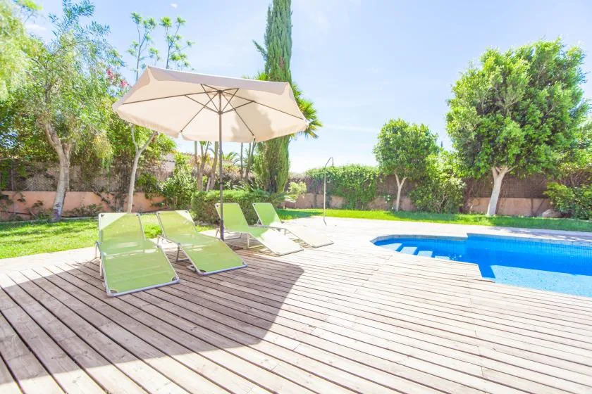 Una piscina junto a una terraza de madera con tumbonas y una sombrilla.