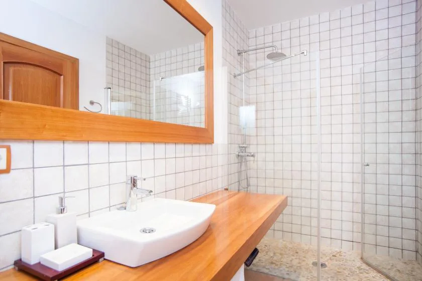 Baño alicatado con lavabo blanco, encimera de madera y mampara de ducha de vidrio.