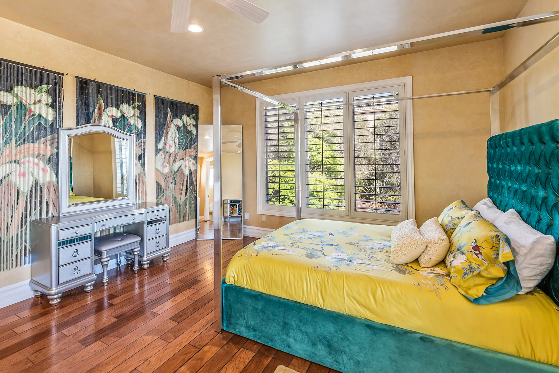 A bedroom with a teal bed, yellow bedding, and large shuttered windows.