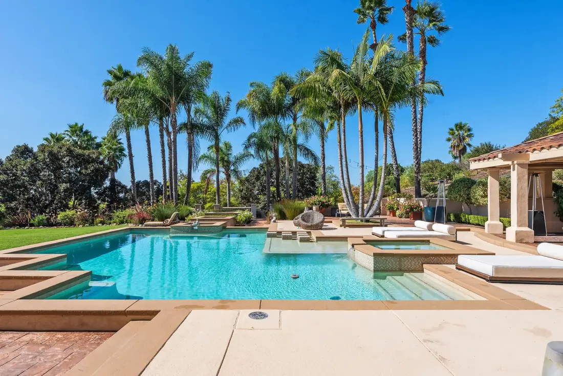 A swimming pool is surrounded by palm trees and a sunny patio.