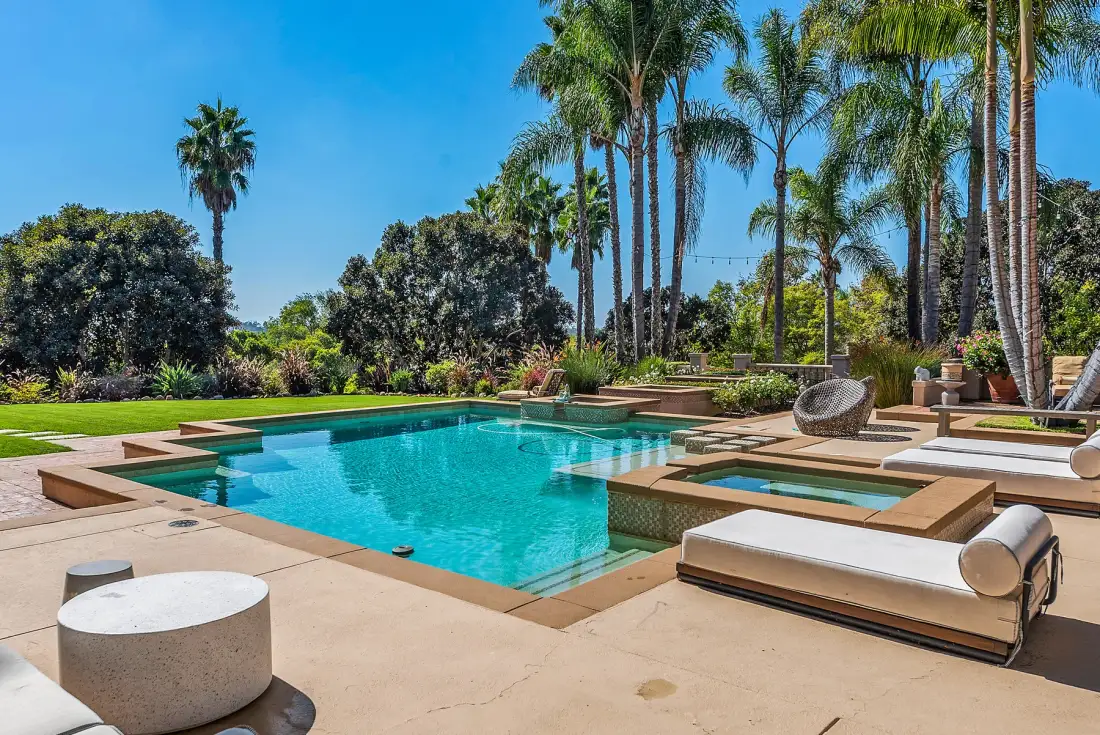 An outdoor swimming pool is surrounded by paved decking, lounge seating, and palm trees.