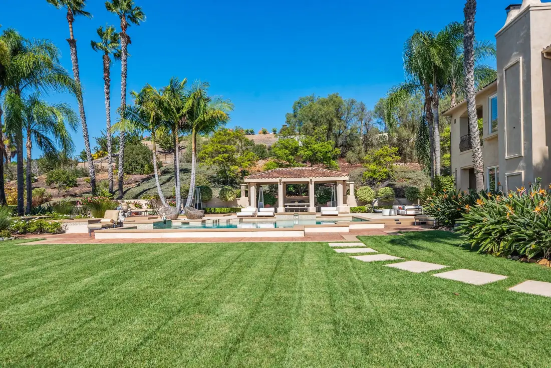 A landscaped outdoor pool area with a pavilion, palm trees, and lawn.