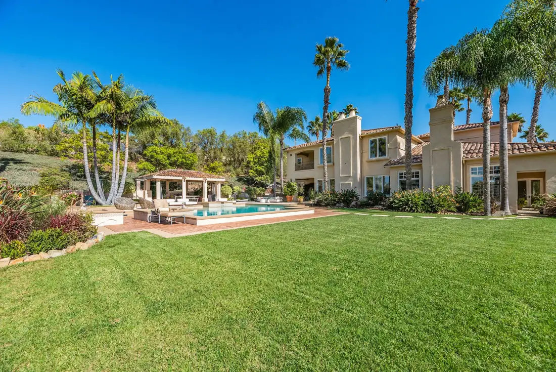 A landscaped lawn leads to a pool and a large house beneath palm trees.