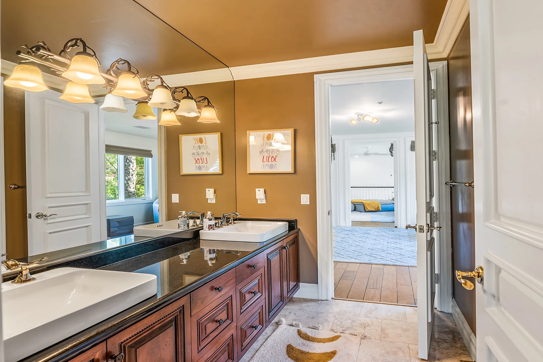A bathroom with a long vanity, wide mirror, and open doorway to a bedroom.